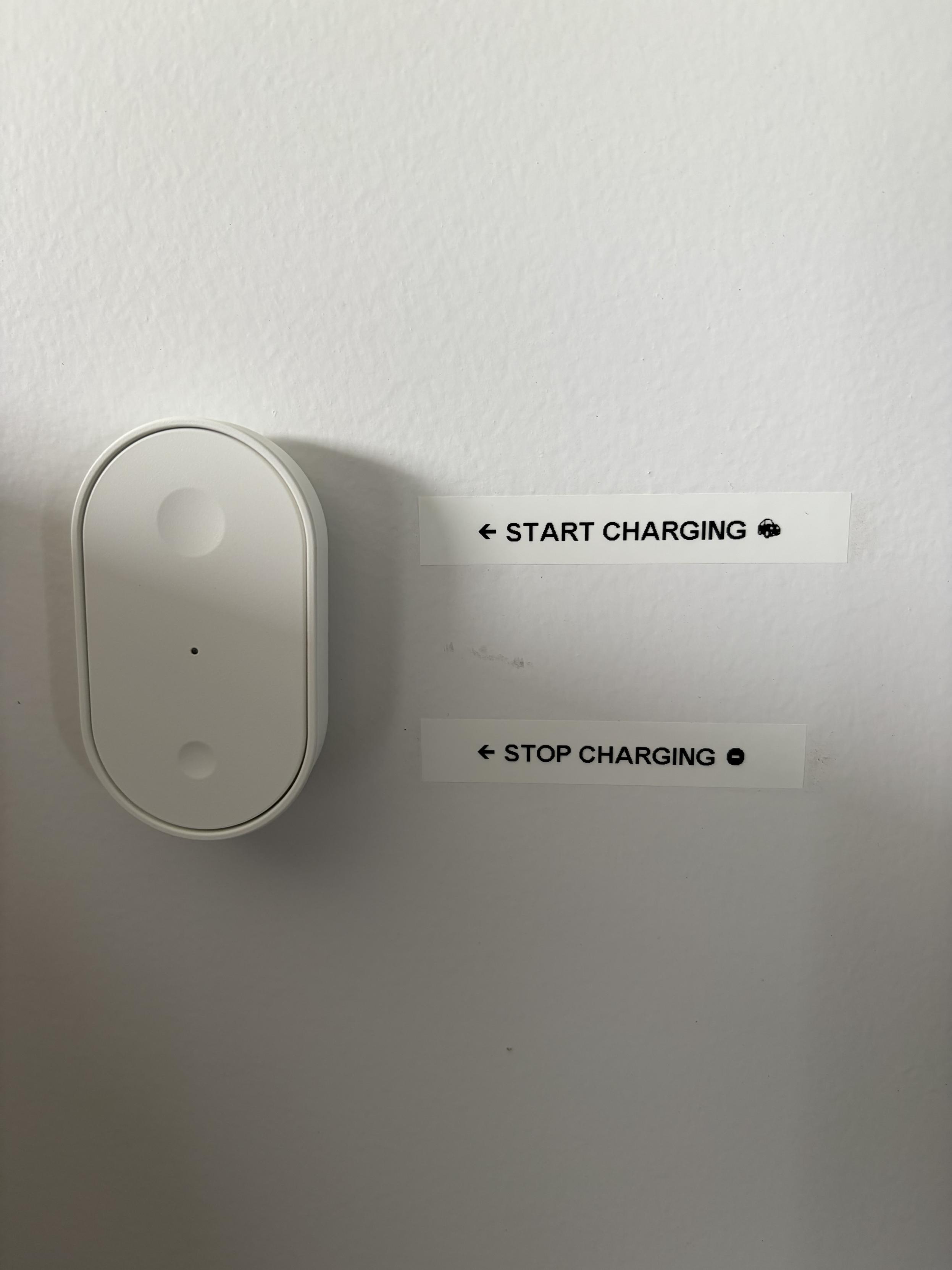 An ikea button remote on a wall, with two stickers. One says to start charging, the other to stop it.